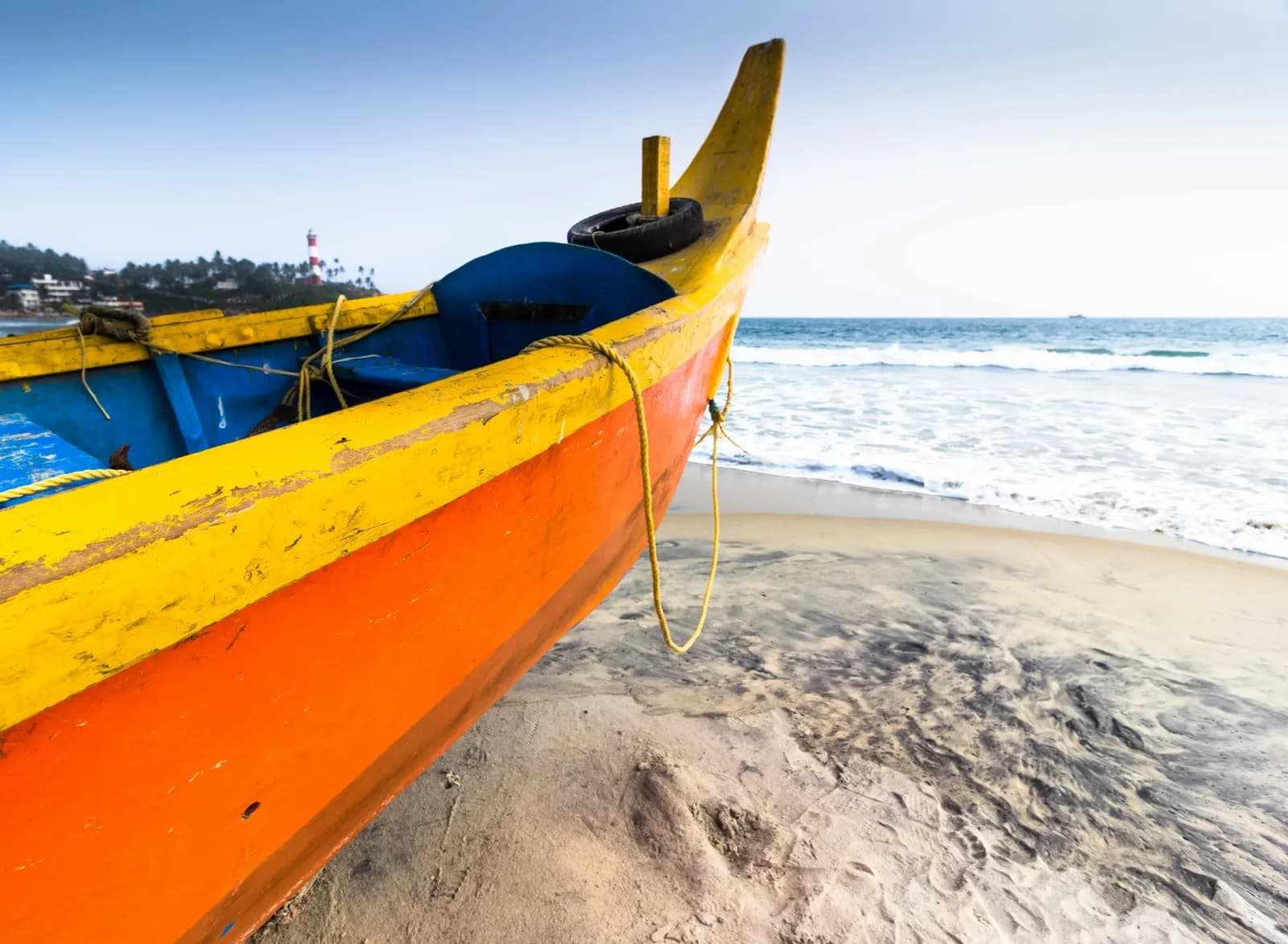 A brightly painted fishing boat on the sand at Kovalam Beach, with the lighthouse in the distance